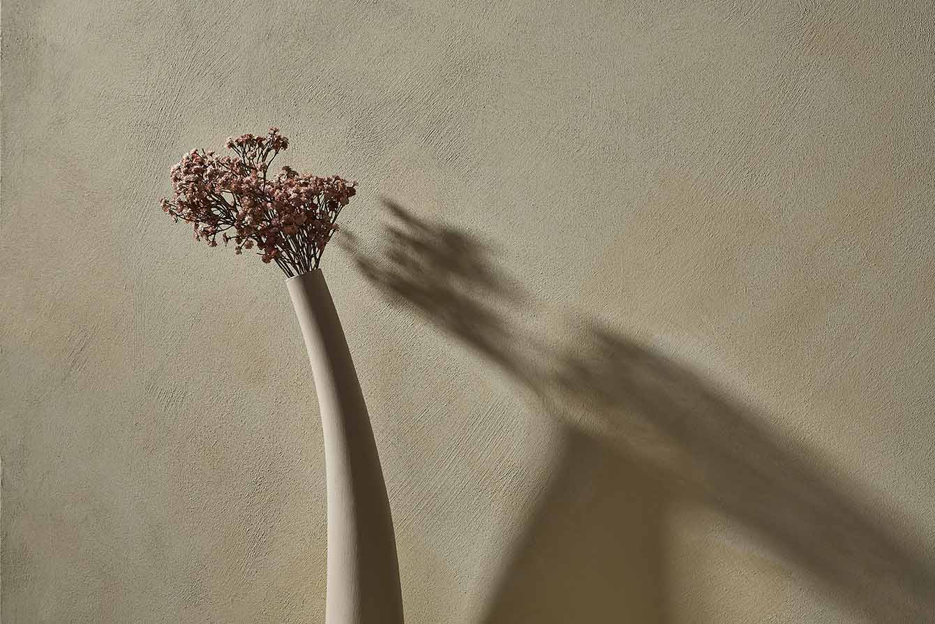 Tall sculptural vase with dried pink flowers casting a dramatic shadow on a textured wall